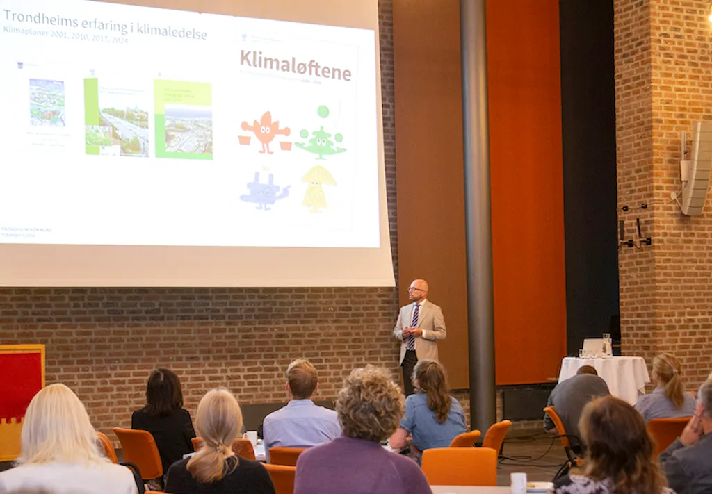 Byrådsleder Kristian Dahlberg Hauge står på scenen og snakker til publikum. På skjermen bak er det en presentasjon som heter Klimaløftene.