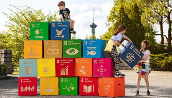 Bilde av alle bærekraftsmålene satt oppå hverandre. En gutt sitter på toppen, mens to jenter løfter opp det siste målet.