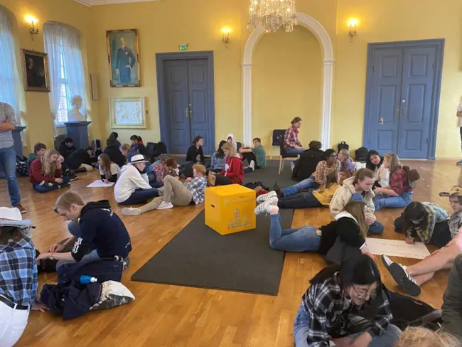 Bilde av vgs-elever som sitter rundt på gulvet og løser oppgaver. Midt i blant dem ser vi en gul kloss med bærekraftsmål 11 på.