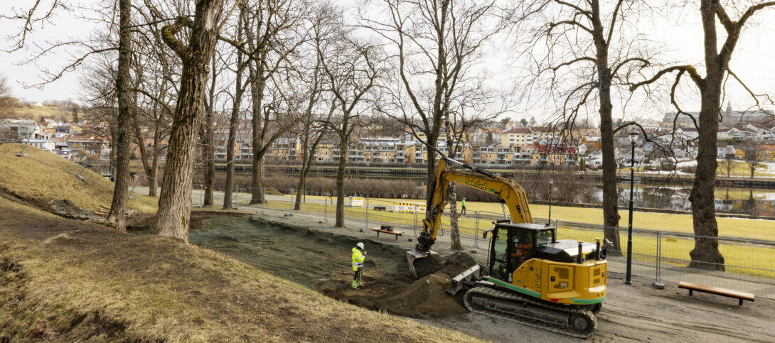 Bilde av en elektrisk drevet gravemaskin som graver på lekeplassområdet på Marinen. Vi ser husrekka på andre siden av Nidelva i bakgrunnen.