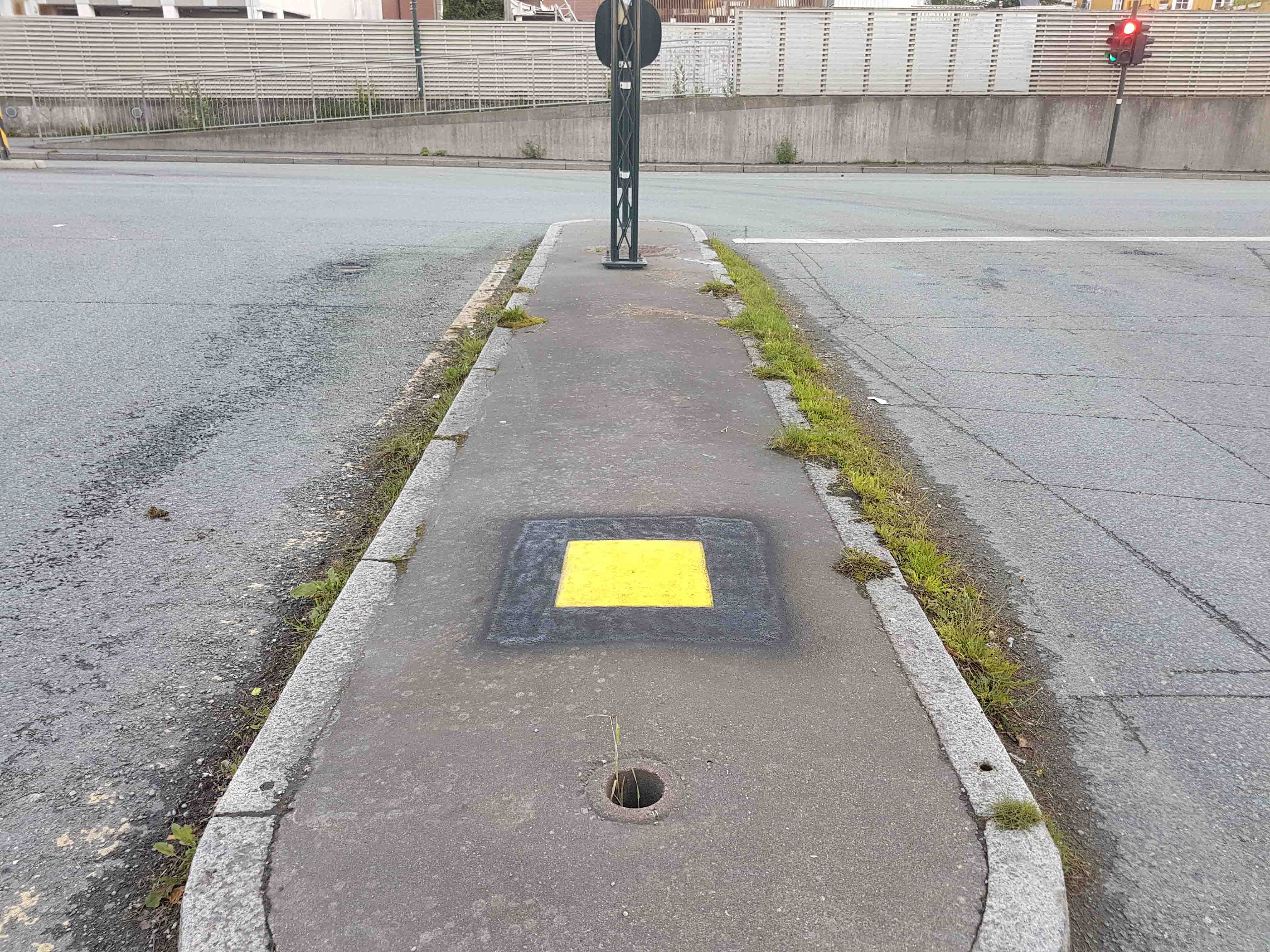 en gul firkant med svart ramme rundt som er malt på asfalten på en midtrabatt.