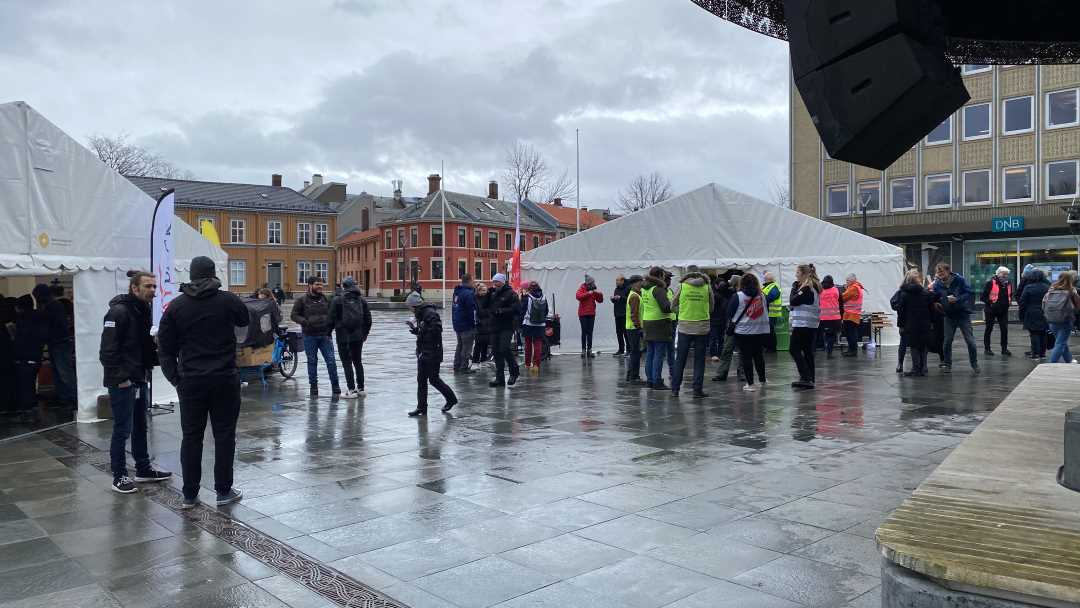 Trondheim Torv på en dag med regn. Flere oppmøtte mennesker foran to telt og scenen.