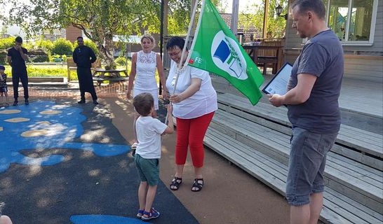 Et barn tar imot Grønt flagg- flagget fra daværende ordfører Rita Ottervik. Flere voksne står rundt dem. Det er sommer og sol.